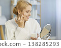 Smiling young woman in bathrobe applying face cream and looking in mirror during skincare routine. 138285293