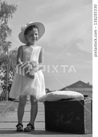 A cute little girl in a white dress with a plush toy and an old suitcase. The adorable smiling girl is on a train platform. The concept of the end of summer vacation, traveling, and a carefree youth. A cute little girl in a white dress with a plush toy and an old suitcase. The adorable smiling girl is on a train platform. The concept of the end of summer vacation, traveling, and a carefree youth. 138285330