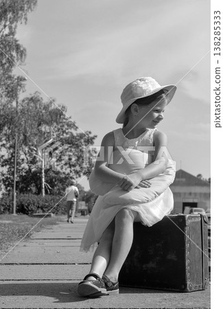 Young girl in a hat sits on a vintage suitcase with a pillow, waiting for a train Young girl in a hat sits on a vintage suitcase with a pillow, waiting for a train 138285333