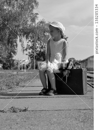Young girl in a white dress and hat sits on a vintage suitcase with a teddy bear Young girl in a white dress and hat sits on a vintage suitcase with a teddy bear 138285334