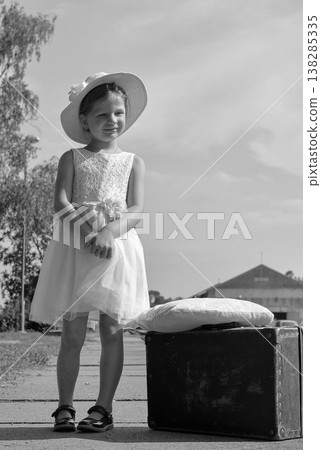 Young girl in a white dress and hat standing next to a vintage suitcase on a sunny day 138285335
