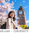 A spring portrait of a woman with a backpack on, smiling and looking up against the backdrop of a clock tower in the background, against a backdrop of a blue sky and cherry blossoms in full bloom. 138285673