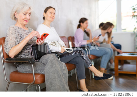 Elderly positive woman communicates on mobile phone while sitting on chair in the company office waiting for specialist Elderly positive woman communicates on mobile phone while sitting on chair in the company office waiting for specialist 138288065