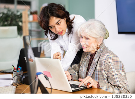 Asian girl leaned over to seated senior woman employee, helps with work and looks at laptop screen 138288330