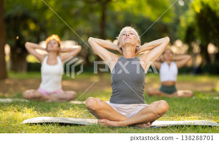 Happy old lady and class of calm sports women in activewear stretching muscles of neck while sitting in lotus pose on mats during yoga training together at city park Happy old lady and class of calm sports women in activewear stretching muscles of neck while sitting in lotus pose on mats during yoga training together at city park 138288701