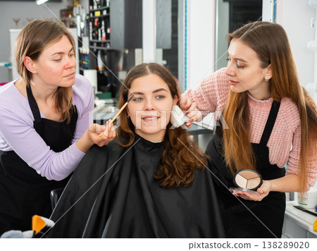 Two makeup artists apply makeup on the face of a young woman Two makeup artists apply makeup on the face of a young woman 138289020
