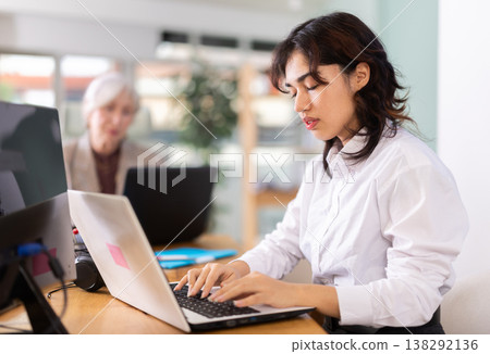 Young woman sitting at desk and typing on laptop in modern office Young woman sitting at desk and typing on laptop in modern office 138292136