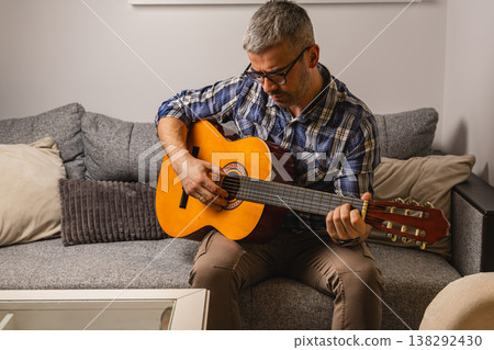 Man playing guitar on a couch in a living room during the day Man playing guitar on a couch in a living room during the day 138292430