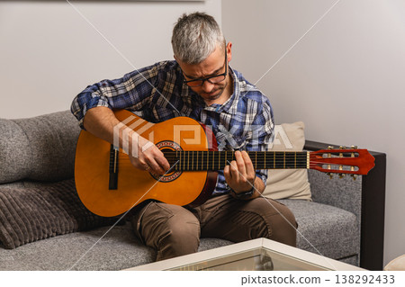Man plays guitar while sitting on a couch in a cozy room Man plays guitar while sitting on a couch in a cozy room 138292433