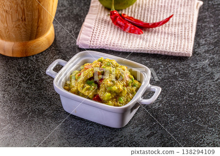 Homemade guacamole dip in a ceramic bowl with fresh avocado, chili peppers, and a wooden mortar on a dark background. 138294109