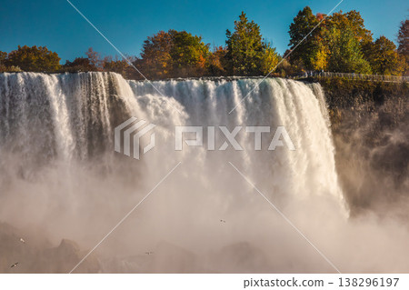 Niagara Falls massive waterfall with mist clouds and autumn trees above cliff Niagara Falls massive waterfall with mist clouds and autumn trees above cliff 138296197