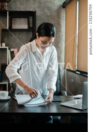 Businesswoman in a white shirt organizing papers at a desk in a contemporary office setting. Businesswoman in a white shirt organizing papers at a desk in a contemporary office setting. 138298704