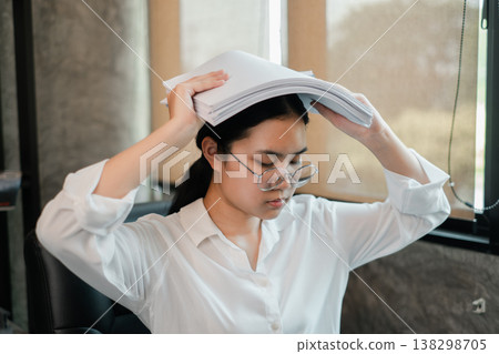 A young woman in an office setting holds a stack of papers on her head, appearing stressed or overwhelmed. A young woman in an office setting holds a stack of papers on her head, appearing stressed or overwhelmed. 138298705