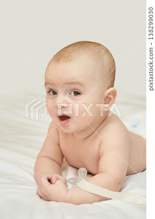 Caucasian Baby Lying On Stomach On White Bed, Wearing Diaper, Expressing Surprise, Indoor Soft Caucasian Baby Lying On Stomach On White Bed, Wearing Diaper, Expressing Surprise, Indoor Soft 138299030
