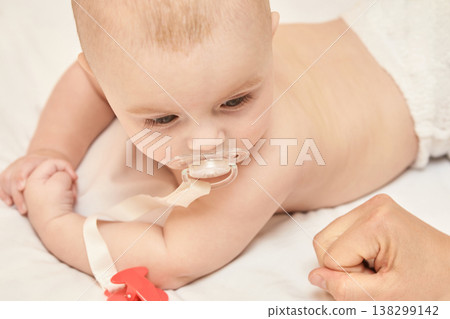Caucasian Male Baby With Pacifier Lying On Stomach In White Diaper On Bed, Arm Fist Close-Up 138299142