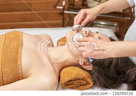 Asian Female Receiving Facial Treatment With Cold Stone Massage At Spa For Relaxation Asian Female Receiving Facial Treatment With Cold Stone Massage At Spa For Relaxation 138299219