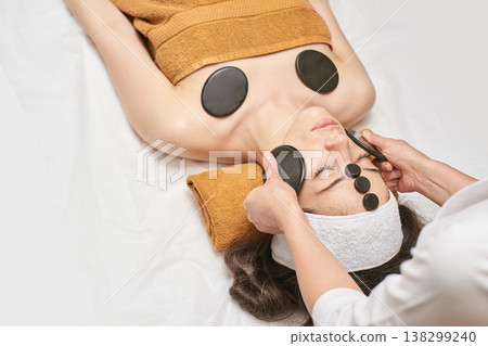 Young Caucasian Female Relaxing During Hot Stone Facial Massage At Spa For Rejuvenation Young Caucasian Female Relaxing During Hot Stone Facial Massage At Spa For Rejuvenation 138299240