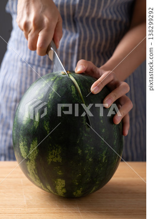 Woman's hands holding a knife and preparing to slice a fresh watermelon on a wooden cutting board 138299292