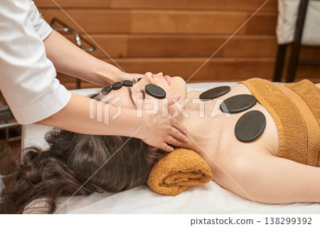 Young Caucasian Female Enjoying Relaxing Hot Stone Massage Therapy In Spa Setting With Therapists 138299392