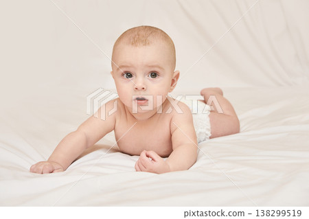 Curious Caucasian Baby Lying On Stomach In Diaper On White Bed Linen Looking At Camera 138299519
