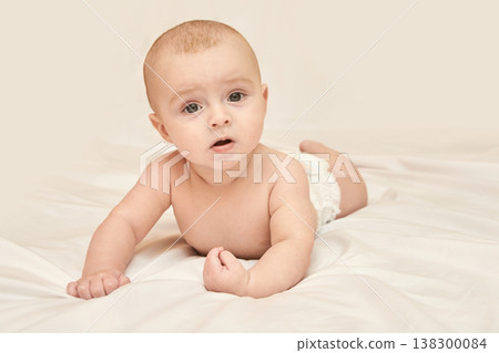 Caucasian Baby In Diaper Lying On Stomach On White Bed With Curious Expression Looking Forward Caucasian Baby In Diaper Lying On Stomach On White Bed With Curious Expression Looking Forward 138300084