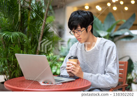 Glasses asian man holding coffee both hands looking at laptop while sitting at table in garden cafe. Glasses asian man holding coffee both hands looking at laptop while sitting at table in garden cafe. 138302202