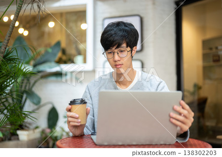 Glasses asian man looking at laptop holding a lid and coffee while sitting at table in garden cafe. Glasses asian man looking at laptop holding a lid and coffee while sitting at table in garden cafe. 138302203
