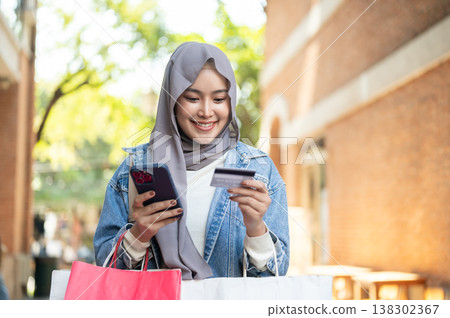 Happy asian muslim woman tourist holding phone looking at credit card while shopping in town market. 138302367