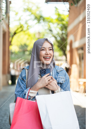 Happy asian muslim woman tourist holding hugging phone carrying shopping bags as walking in market. Happy asian muslim woman tourist holding hugging phone carrying shopping bags as walking in market. 138302409