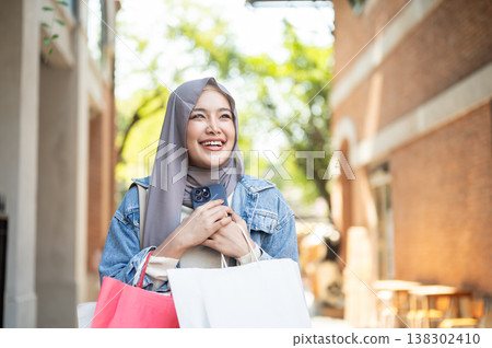 Smiling asian muslim woman tourist holding hugging phone carrying shopping bags as walking in market Smiling asian muslim woman tourist holding hugging phone carrying shopping bags as walking in market 138302410