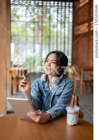 Pretty asian woman holding pen looking over tablet aside coffee on wooden table as sitting in cafe. Pretty asian woman holding pen looking over tablet aside coffee on wooden table as sitting in cafe. 138303046