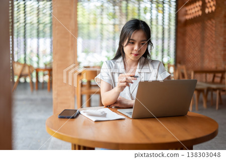 Pretty asian woman holding pen pointing and looking at laptop while sitting at wooden table in cafe. Pretty asian woman holding pen pointing and looking at laptop while sitting at wooden table in cafe. 138303048