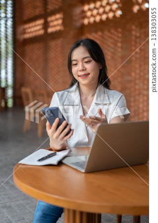 Pretty asian woman holding and looking at phone over book and laptop sitting at wooden table in cafe Pretty asian woman holding and looking at phone over book and laptop sitting at wooden table in cafe 138303055