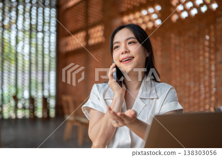 Pretty asian woman holding phone talking on call over laptop while sitting in wooden pavilion cafe. 138303056