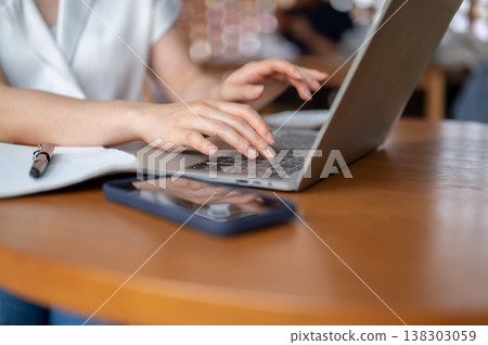 Close up of woman's hand typing on laptop aside book and phone while sitting at wooden table in cafe Close up of woman's hand typing on laptop aside book and phone while sitting at wooden table in cafe 138303059
