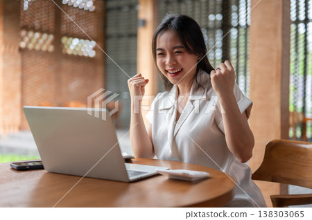 Happy asian woman looking at laptop making a fist in excited while sitting at wooden table in cafe. Happy asian woman looking at laptop making a fist in excited while sitting at wooden table in cafe. 138303065