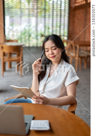 Pretty asian woman holding pen looking at book thinking aside laptop on table while sitting in cafe. 138303075