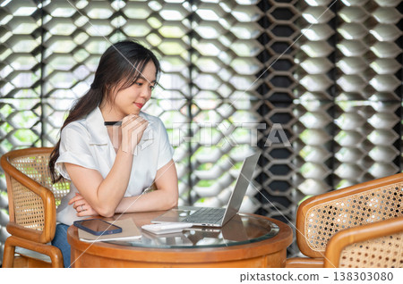 Pretty asian woman holding a pen looking at laptop aside book while sitting at wooden table in cafe. Pretty asian woman holding a pen looking at laptop aside book while sitting at wooden table in cafe. 138303080