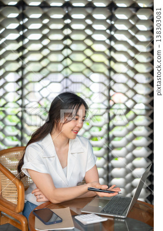 Pretty asian woman holding pen looking at laptop aside book and calculator sitting at table in cafe. Pretty asian woman holding pen looking at laptop aside book and calculator sitting at table in cafe. 138303081