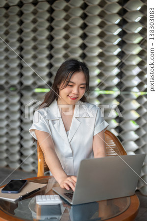 Pretty asian woman typing on laptop aside book and calculator while sitting at glass table in cafe. 138303083