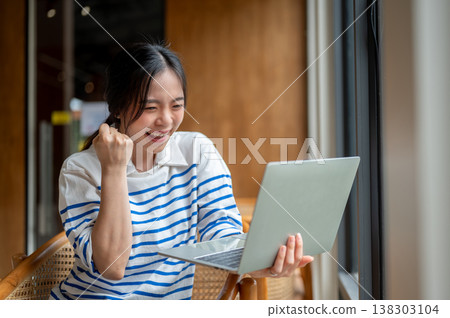 Excited asian woman looking at laptop making a fist while sitting on wooden chair by window in cafe. Excited asian woman looking at laptop making a fist while sitting on wooden chair by window in cafe. 138303104