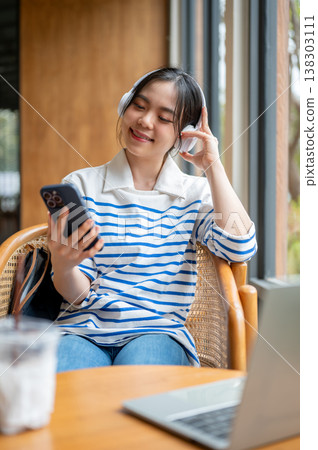 Happy asian woman enjoy listening to music in headphones sitting with laptop at wooden table in cafe Happy asian woman enjoy listening to music in headphones sitting with laptop at wooden table in cafe 138303111