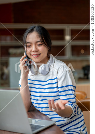 Young asian woman talking on phone looking at laptop while sitting at wooden counter table in a cafe Young asian woman talking on phone looking at laptop while sitting at wooden counter table in a cafe 138303120