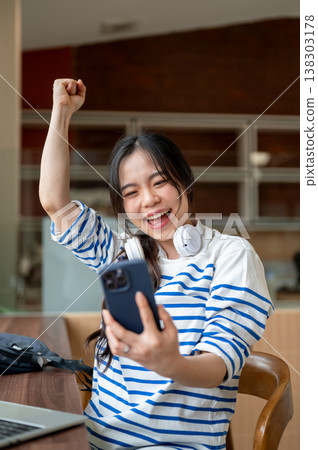 Excited asian woman making raising fist looking at phone while sitting with laptop at table in cafe. 138303178