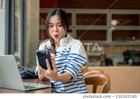 Surprised asian woman pointing looking at phone over laptop while sitting at wooden counter in cafe. Surprised asian woman pointing looking at phone over laptop while sitting at wooden counter in cafe. 138303180