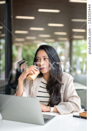 Asian woman holding drinking coffee aside laptop and book as sitting at table in university building Asian woman holding drinking coffee aside laptop and book as sitting at table in university building 138303444