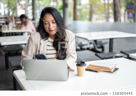 Asian woman looking at phone over laptop aside books and coffee sits at table in university building Asian woman looking at phone over laptop aside books and coffee sits at table in university building 138303445