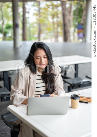 Asian woman looking at phone over laptop aside coffee and book sits at table in university building. Asian woman looking at phone over laptop aside coffee and book sits at table in university building. 138303446