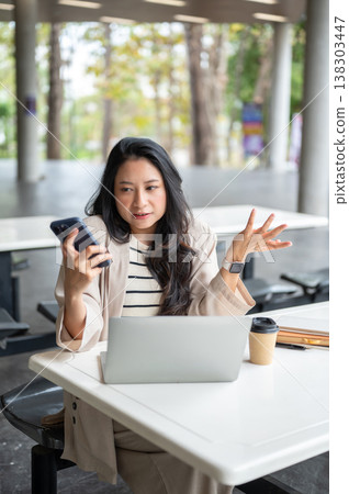 Asian woman shrugging shoulders looking at phone over laptop sitting at table in university building Asian woman shrugging shoulders looking at phone over laptop sitting at table in university building 138303447
