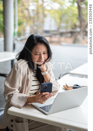 Asian woman frowning and looking at phone over laptop while sitting at table in university building. 138303448
